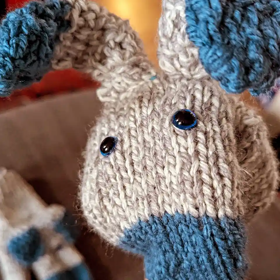 A close-up photo of a hand-knitted stuffed animal, likely a rabbit or similar creature, with a light gray body, blue ears…
