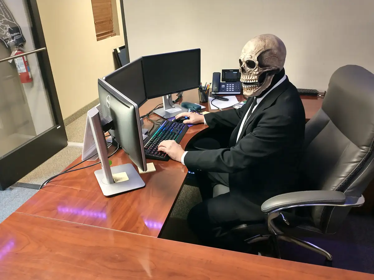 A person wearing a realistic skull mask sits at a wooden office desk, typing on a keyboard with RGB lighting while using two…
