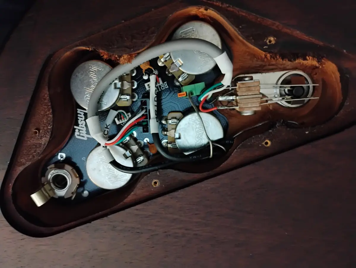 A close-up of the exposed electronics inside a Gibson guitar, showing a circuit board, potentiometers, wiring, and three…
