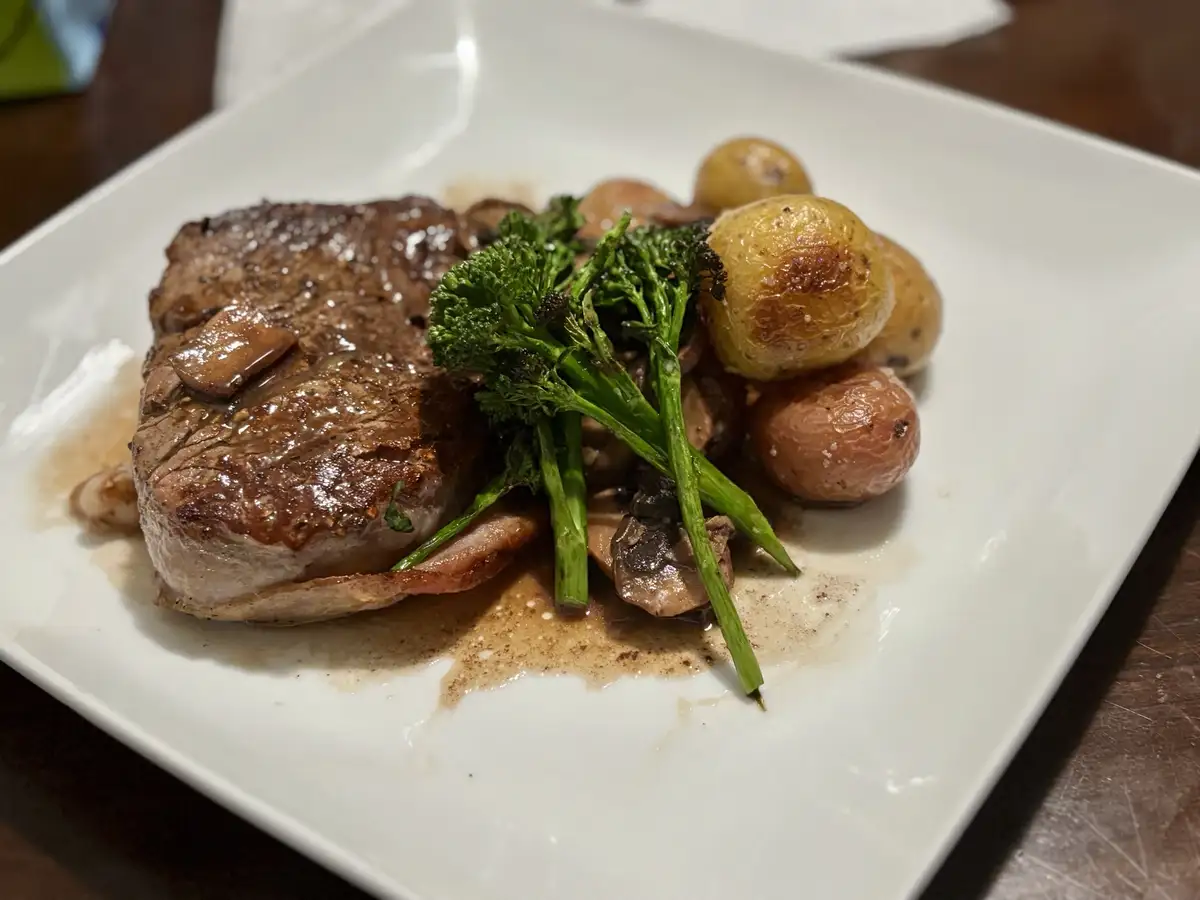 A close-up photo of a plated meal featuring a seared steak, roasted baby potatoes, and broccolini on a white square plate…