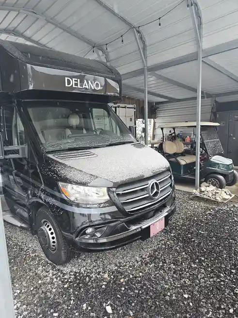 A black Mercedes-Benz motorhome named 'DELANO' is parked under a metal carport, covered in a light layer of snow. In the…
