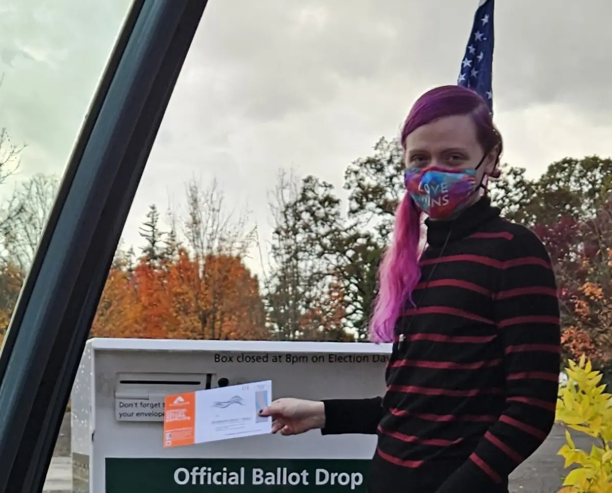 A person with purple hair and a 'Love Wins' face mask stands beside a car, holding a ballot and about to drop it into an…