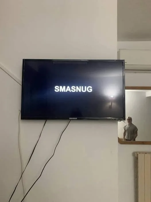 A black Samsung TV mounted on a white wall displays the word 'SMASNUG' in white text. To the right, a man in a cap and shirt…