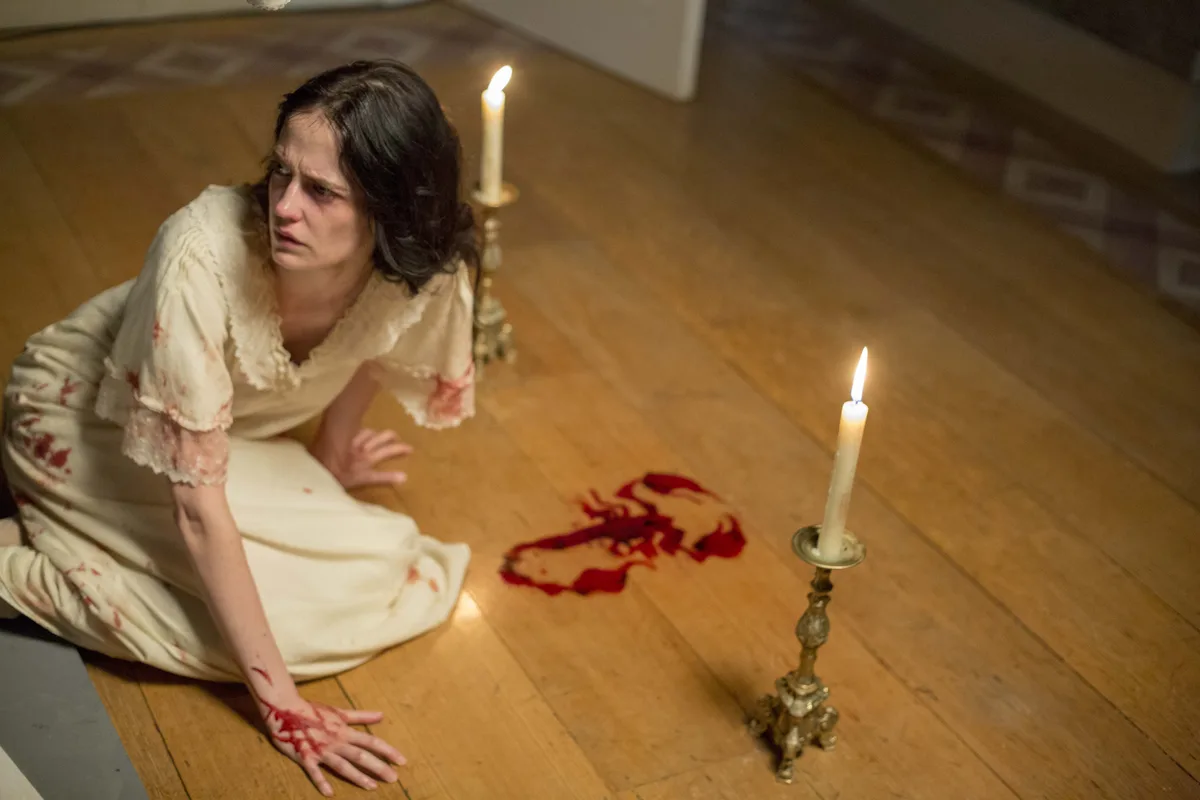 A woman in a blood-stained white nightgown kneels on a wooden floor, looking horrified at a bloodstain shaped like a…