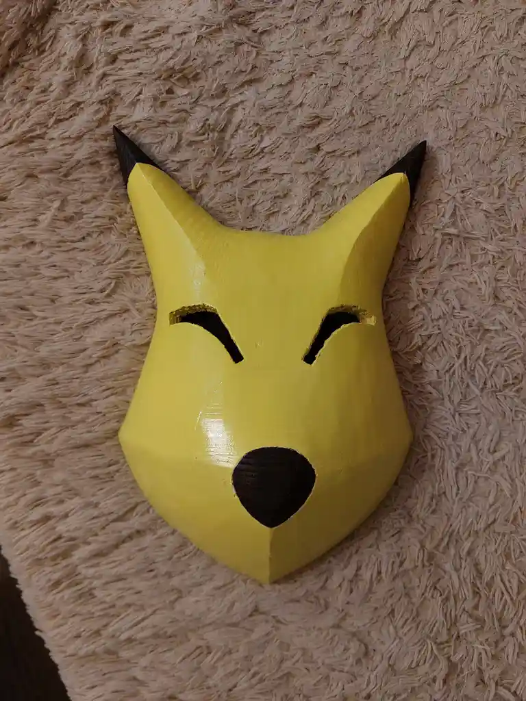 A handmade, yellow, geometrically stylized Pikachu mask with black-tipped ears and a black nose, resting on a beige shaggy…