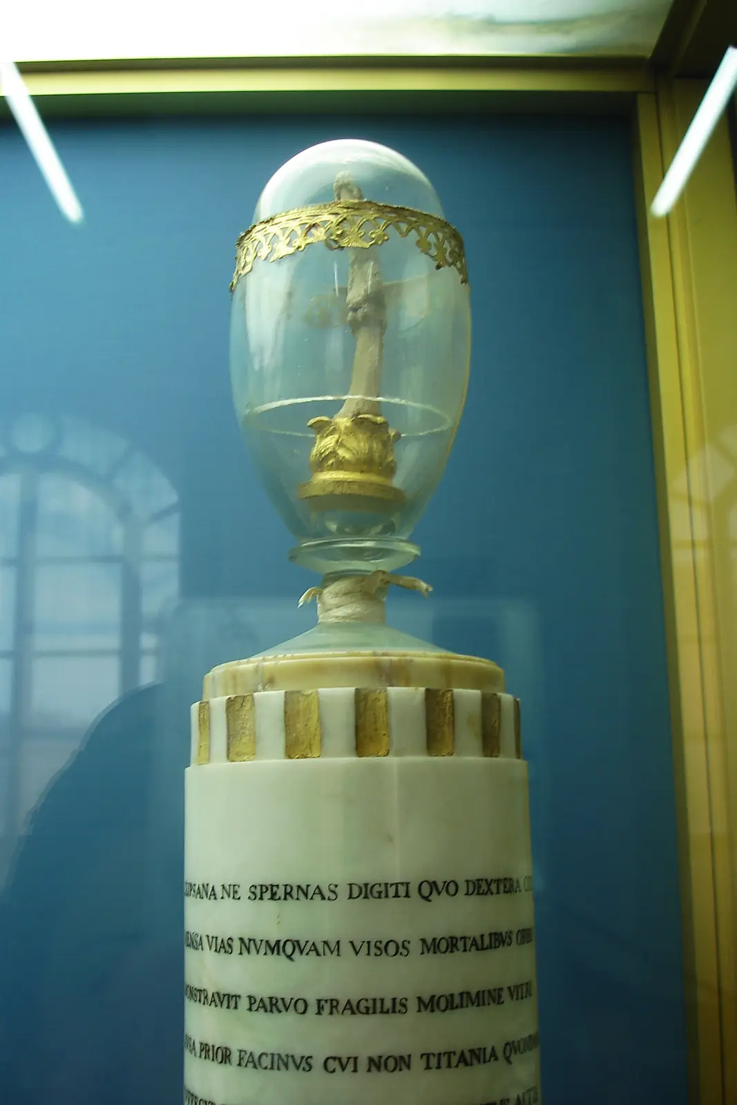 A religious relic, possibly a fragment of the True Cross, is displayed under a glass dome atop a marble pedestal with Latin…