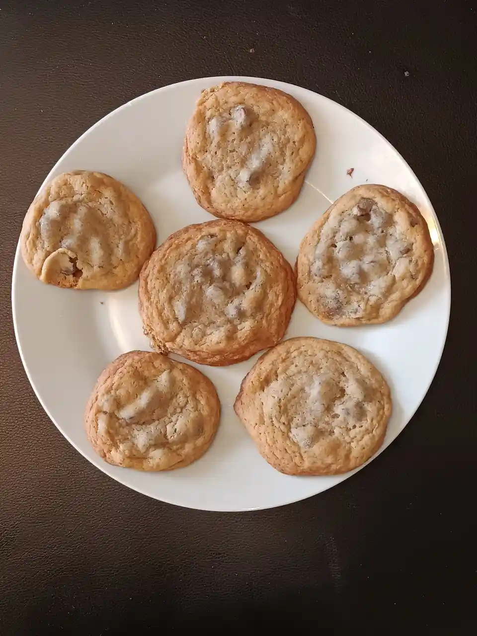 A white plate holds six golden-brown cookies with visible chunks of chocolate or caramel, arranged on a dark brown textured…
