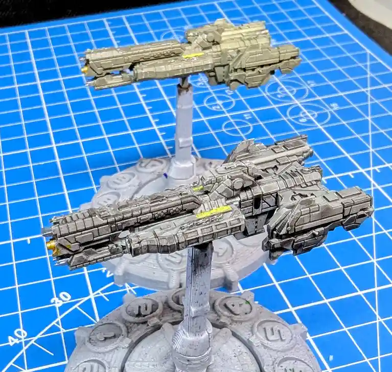 Two intricately detailed science fiction spaceship miniatures are mounted on stands against a blue grid mat. The ship in the…