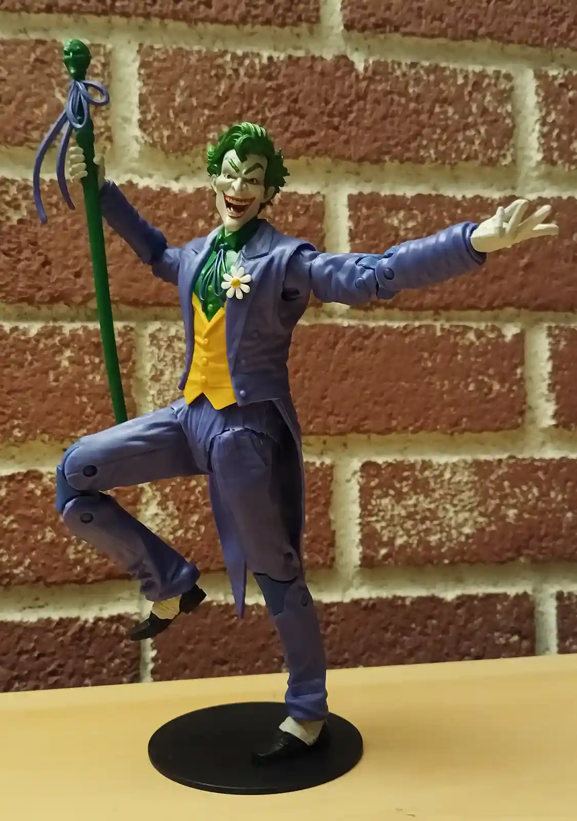A Joker action figure from Batman, posed with arms outstretched and one leg lifted, holding a green staff with a blue…