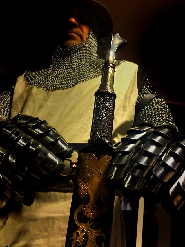 A person in full medieval knight armor, including chainmail coif, plate gauntlets, and a white tunic, holds an ornate sword…