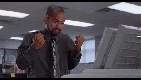 A man in a grey shirt and tie stands in an office with a computer monitor, his fists clenched and face contorted in rage…