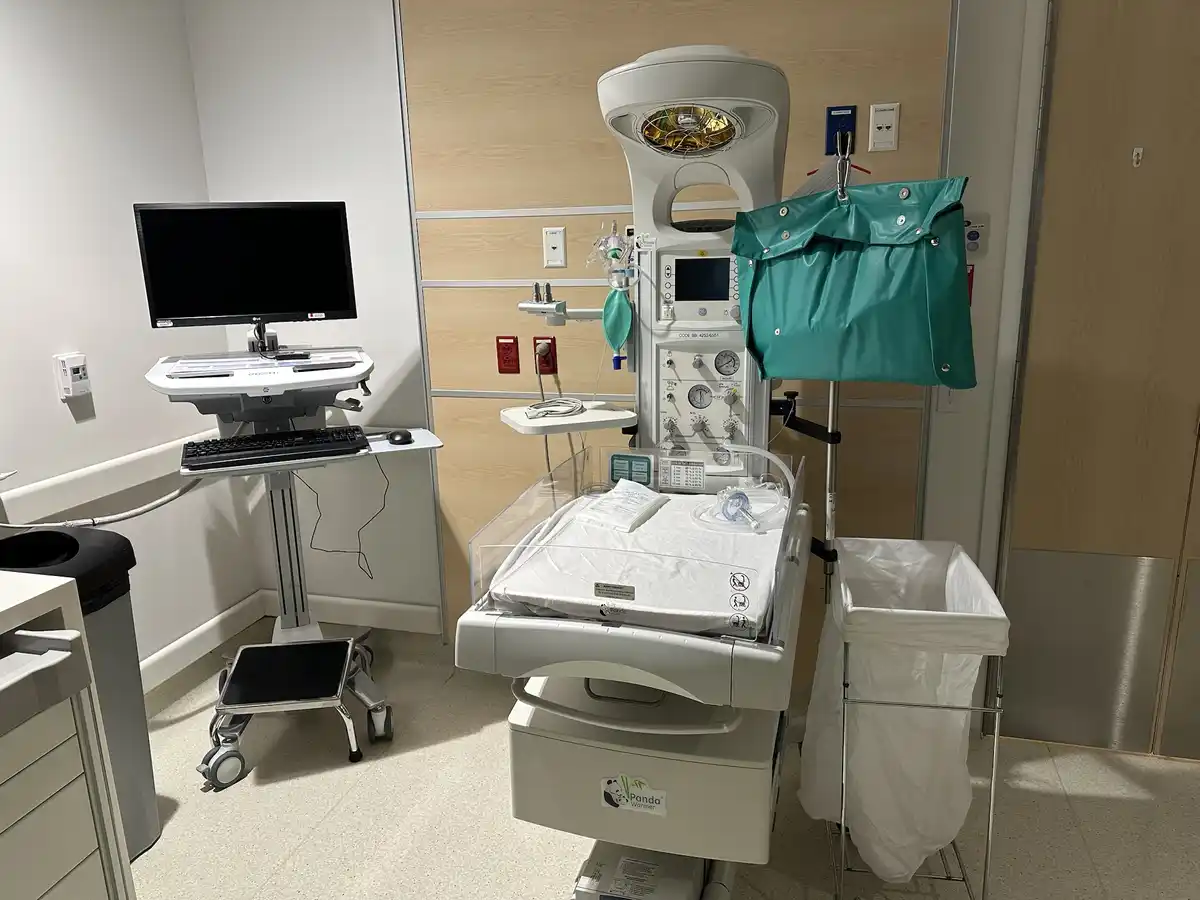 An empty neonatal intensive care unit (NICU) incubator with medical equipment, a computer station, and a trash can in a…