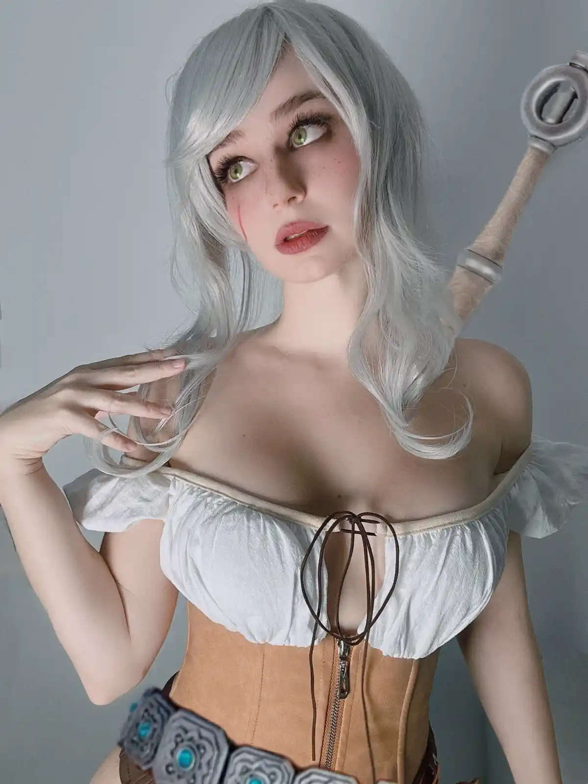 A person with silver hair and green eyes, wearing a white off-the-shoulder top and a tan corset, holds a sword over their…