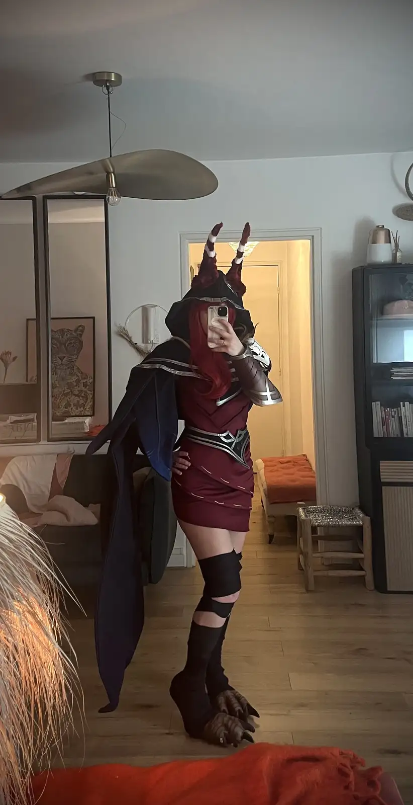 A person in a detailed demon cosplay with red hair, horns, and clawed feet takes a mirror selfie in a living room, holding a…