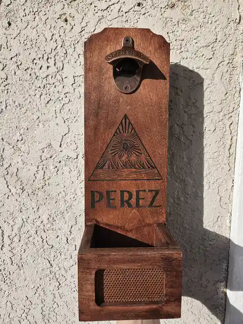 A wooden wall-mounted bottle opener with a built-in tray and perforated speaker grille, featuring an engraved…