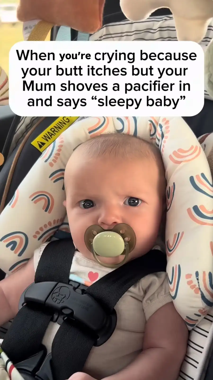 A close-up photo of a baby with wide, expressive eyes looking directly at the camera, wearing a pacifier in their mouth and…