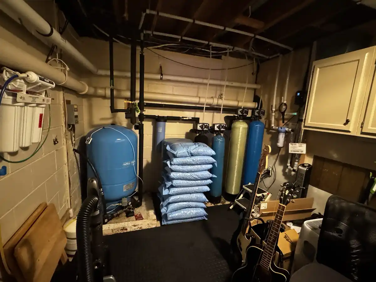 A cluttered basement utility room filled with water filtration equipment, including large blue and green tanks, stacked bags…