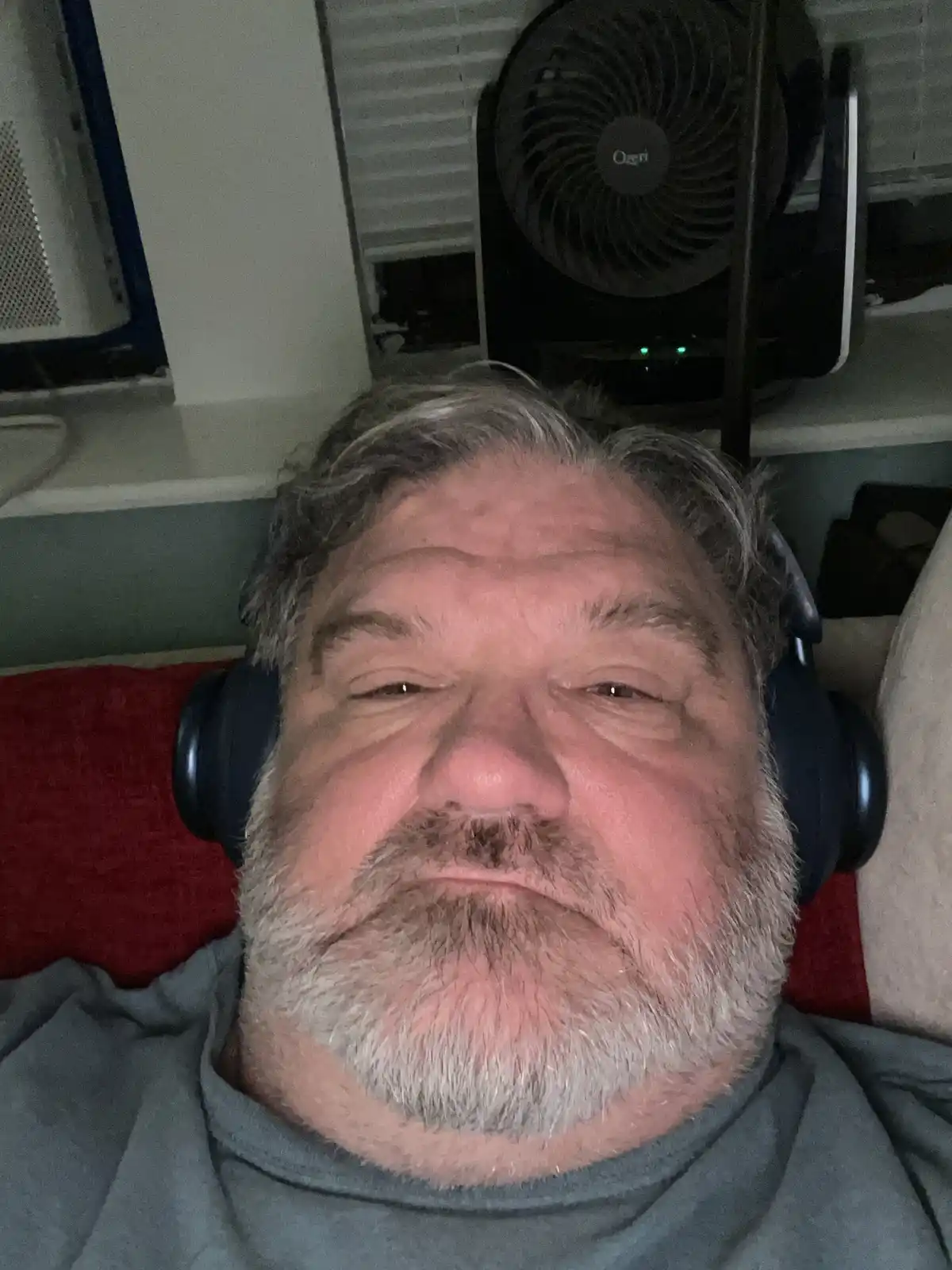 A man with gray hair and a beard, wearing large black headphones, lies back with his eyes half-closed in a relaxed or sleepy…