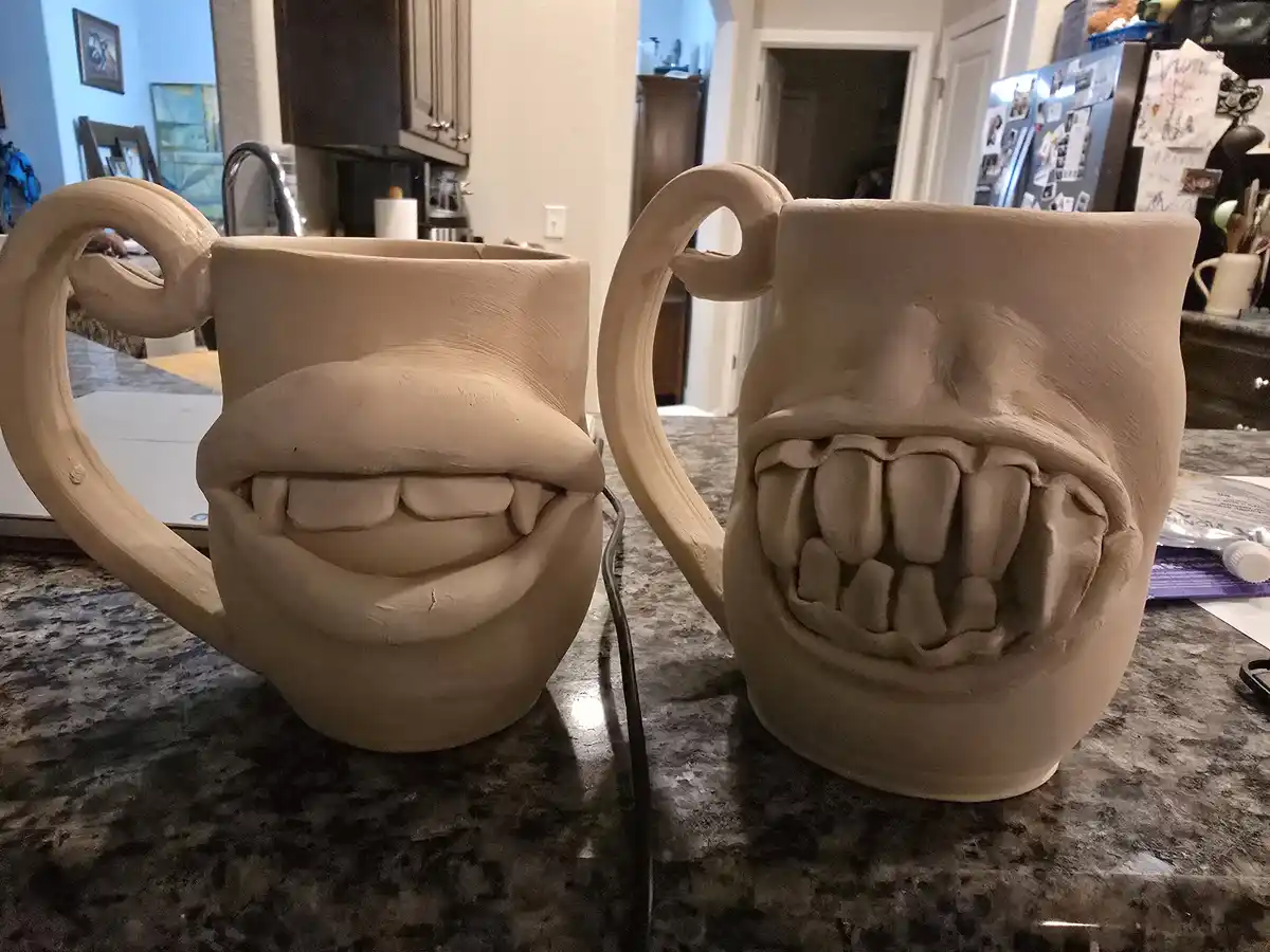 Two handmade clay mugs sit on a kitchen counter, each sculpted with a wide, toothy grin. The mug on the left has a subtle…