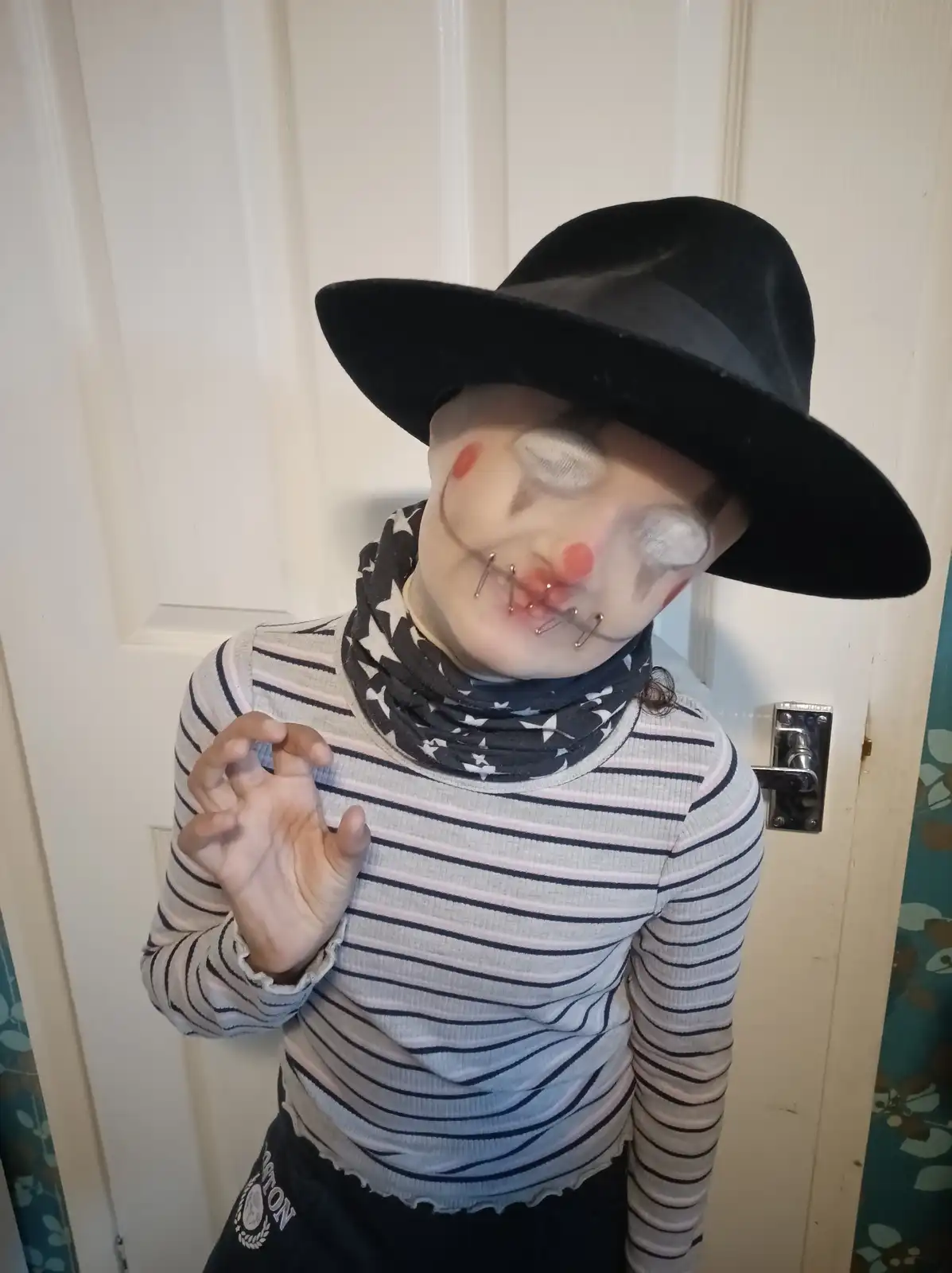 A person dressed as a clown with white face paint, red nose, and red cheeks, wearing a black hat and a striped shirt. Their…