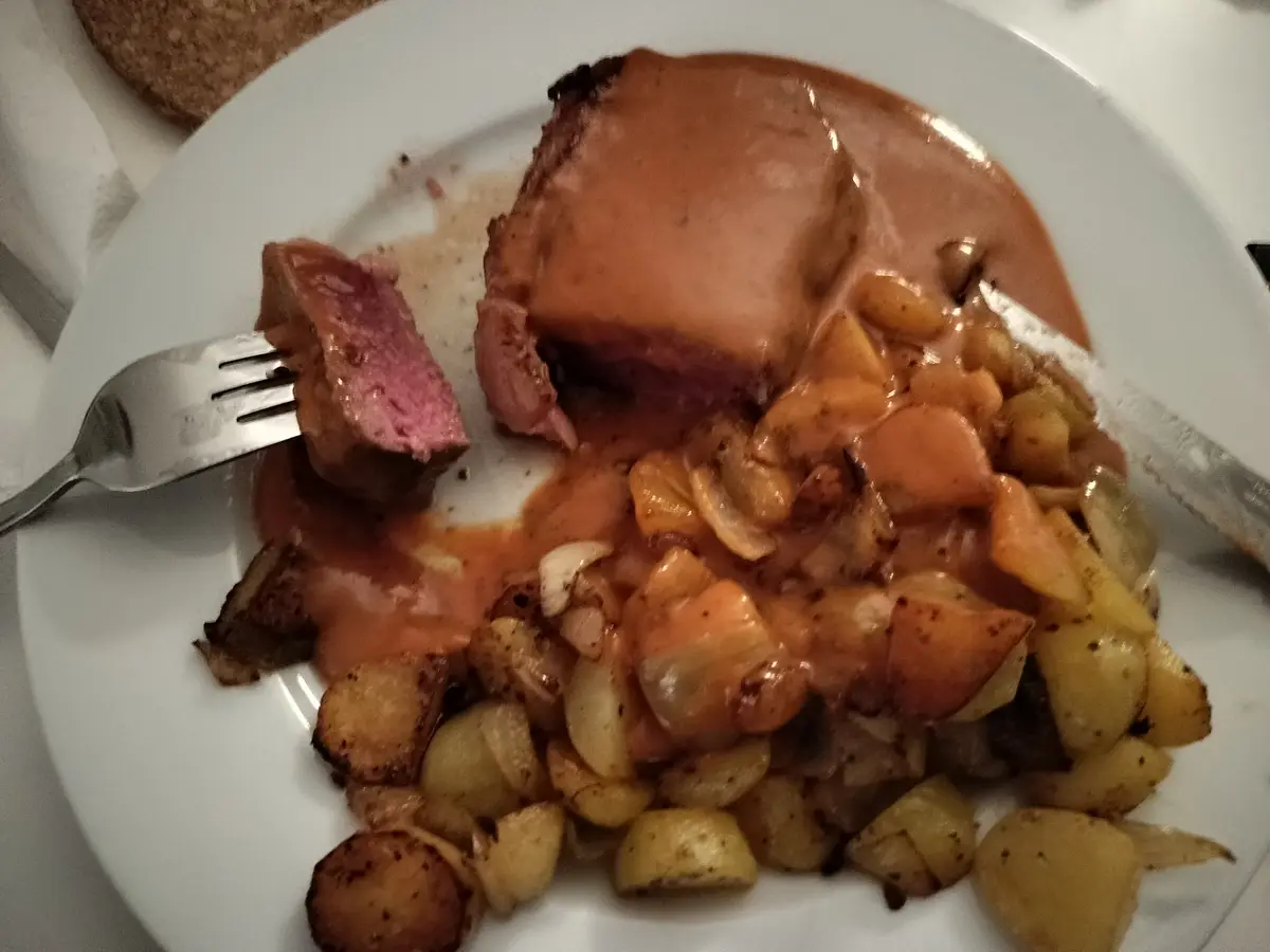 A close-up photo of a partially eaten plate of food featuring a large piece of medium-rare meat covered in sauce, alongside…