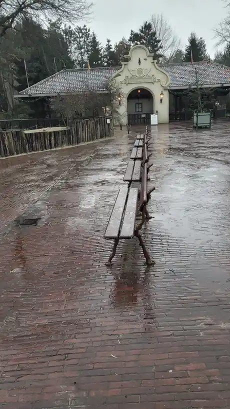 A row of empty, wet benches lines a brick pathway leading to a building entrance labeled 'De Oude Tufferbaan'. The ground is…