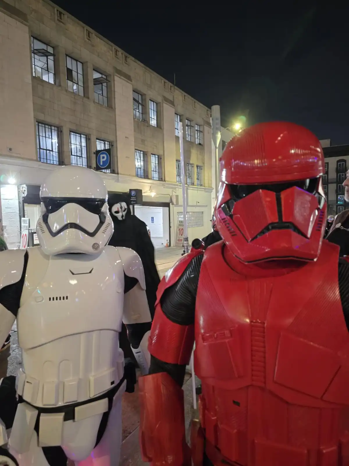 Two people in Star Wars stormtrooper costumes stand side by side at night, one in white armor and the other in red armor…