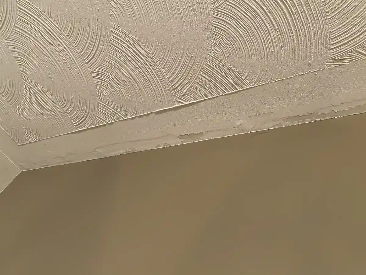 A close-up photo shows a ceiling with an ornate, textured pattern of concentric arcs in beige paint, meeting a plain wall…