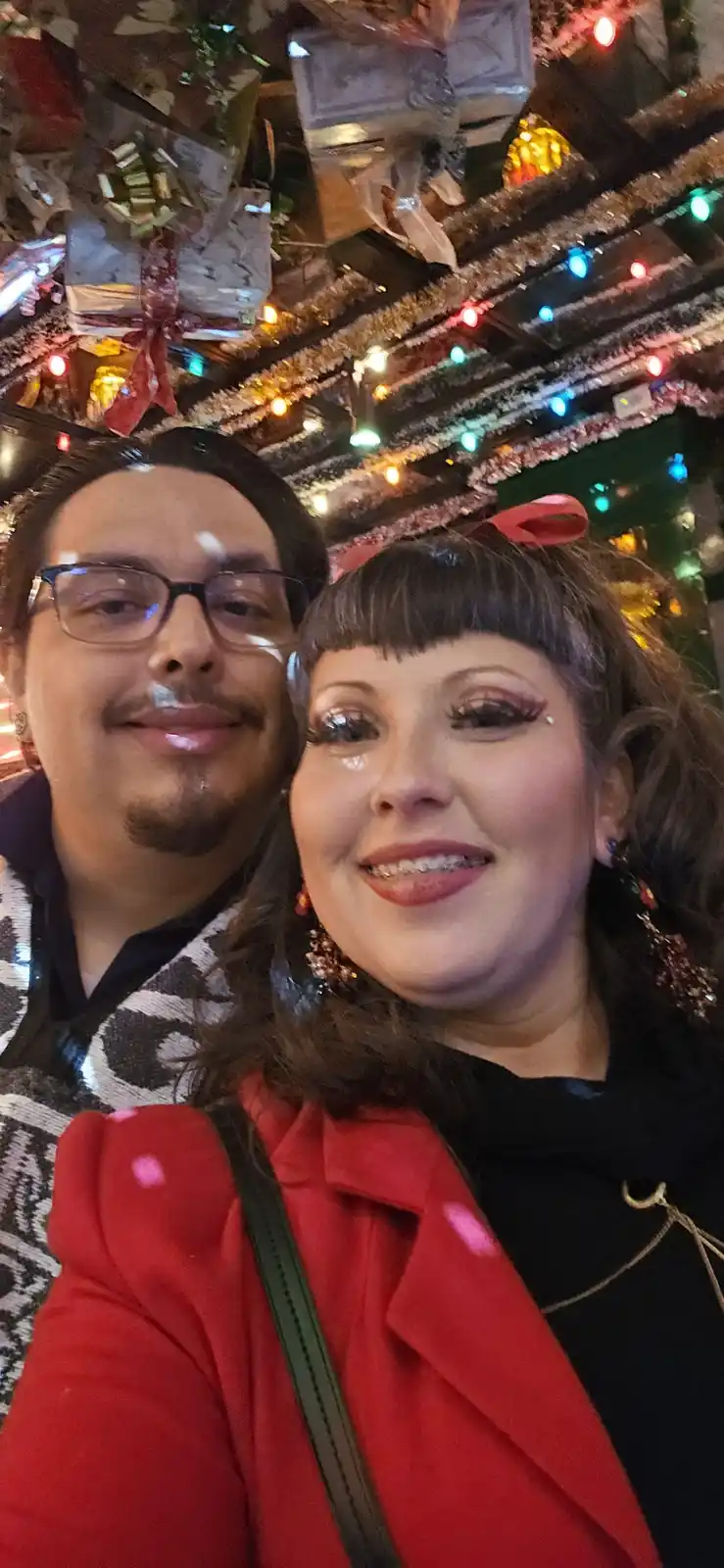 A selfie of a smiling man and woman in a festive, decorated room with lights and wrapped gifts hanging from the ceiling. The…