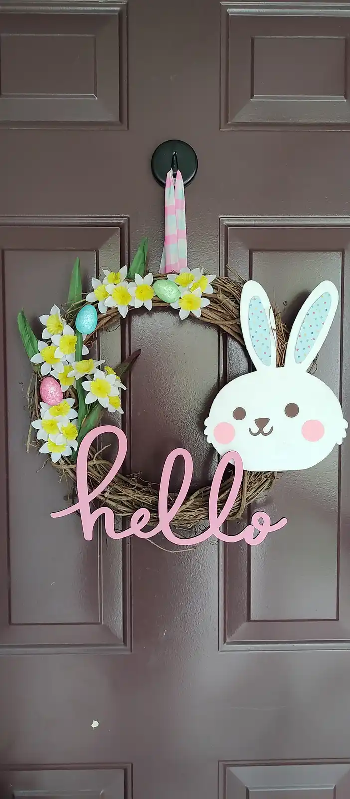 An easter-themed wreath hangs on a dark brown door, featuring a woven base, white daffodils with yellow centers…