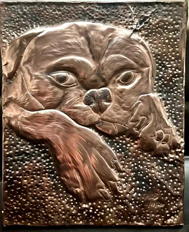 A detailed, hammered copper relief sculpture depicts a dog's face and front paws resting on a textured background. The…
