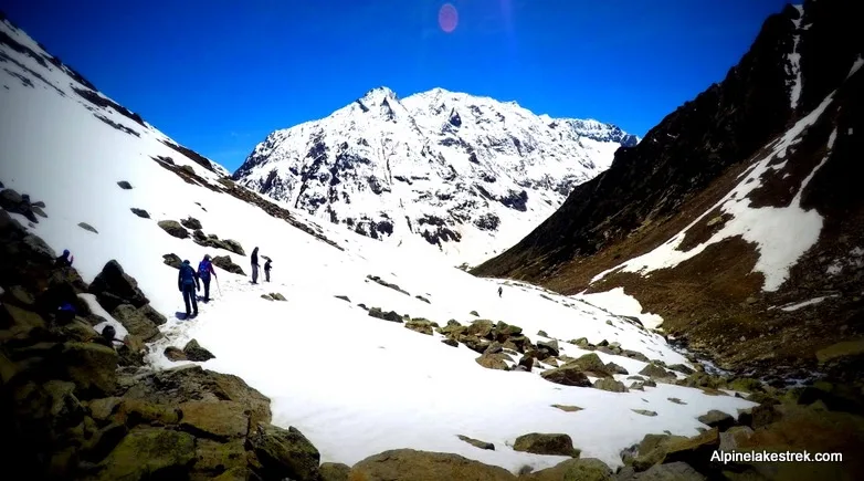 A wide-angle photo shows several hikers trekking across a snowy mountain valley under a clear blue sky, with a massive…