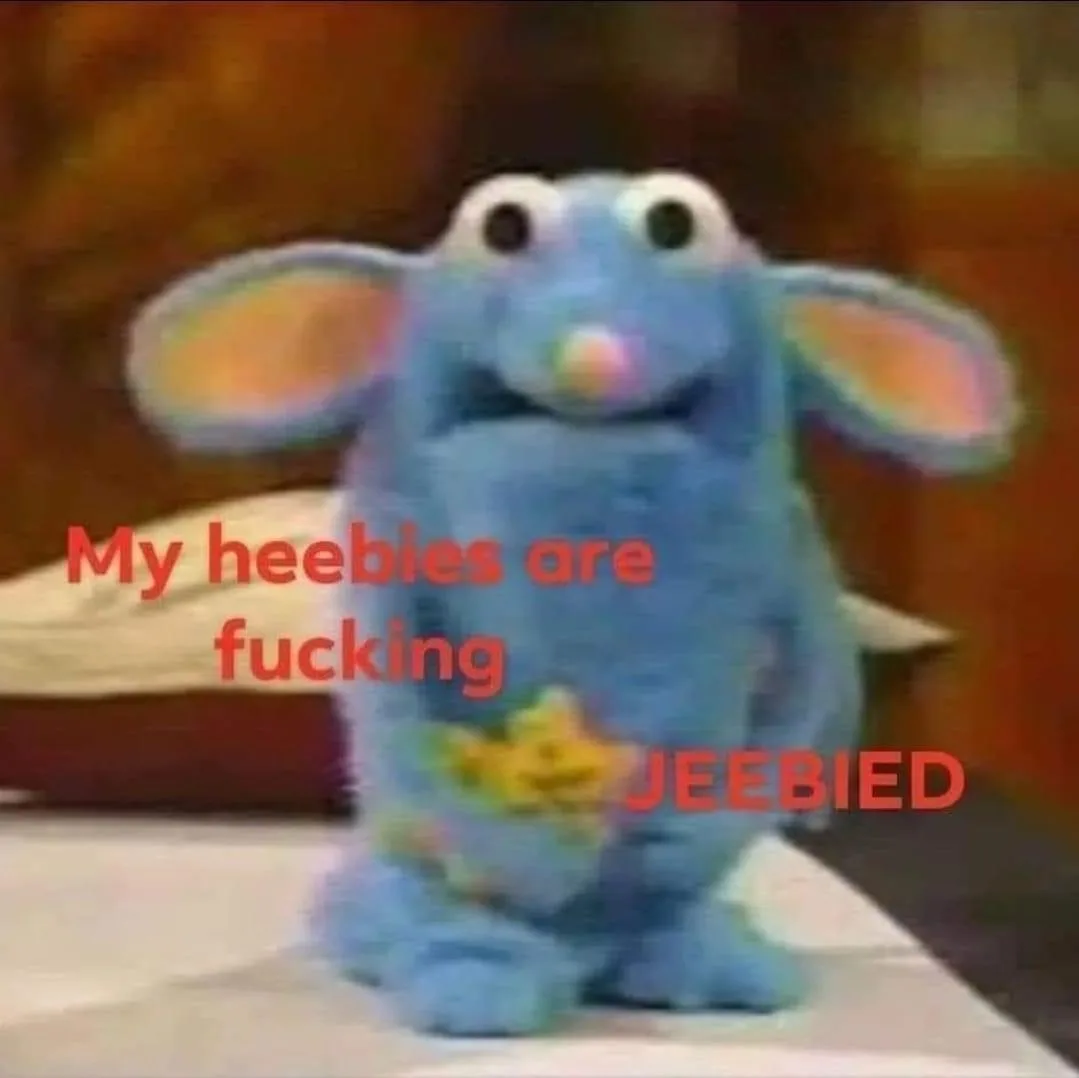 A blurry image of a blue, furry puppet with large ears and a yellow star on its chest, sitting on a white surface. Red text…