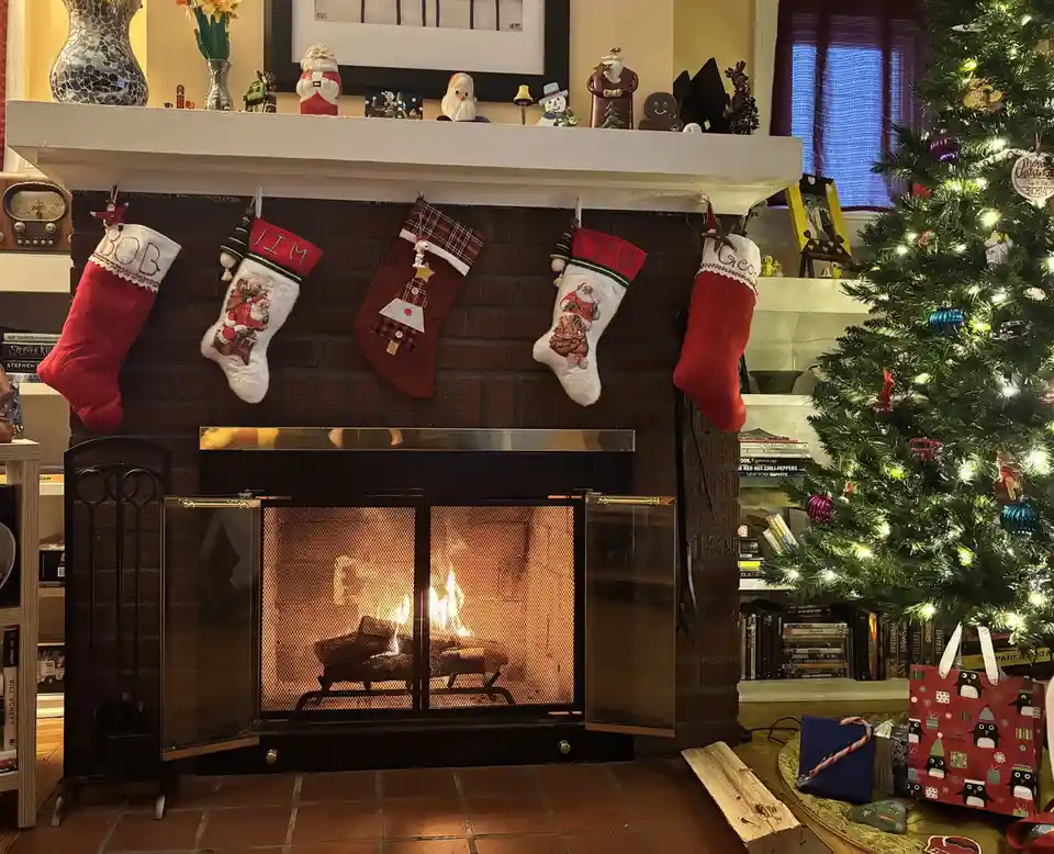 A cozy, decorated living room features a brick fireplace with a fire burning inside, flanked by a lit Christmas tree with…