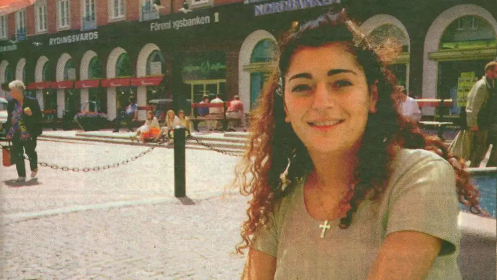 A woman with curly hair and a cross necklace smiles at the camera in a public square. Behind her, a building has signs for…