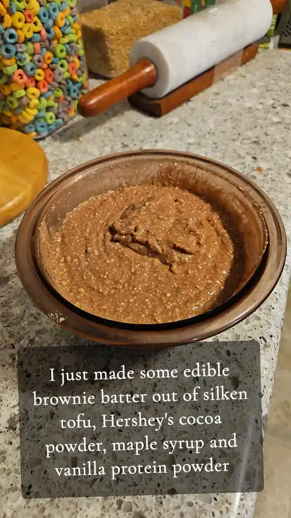 A brown bowl filled with thick, textured brown batter sits on a speckled kitchen counter. In the background, a jar of…