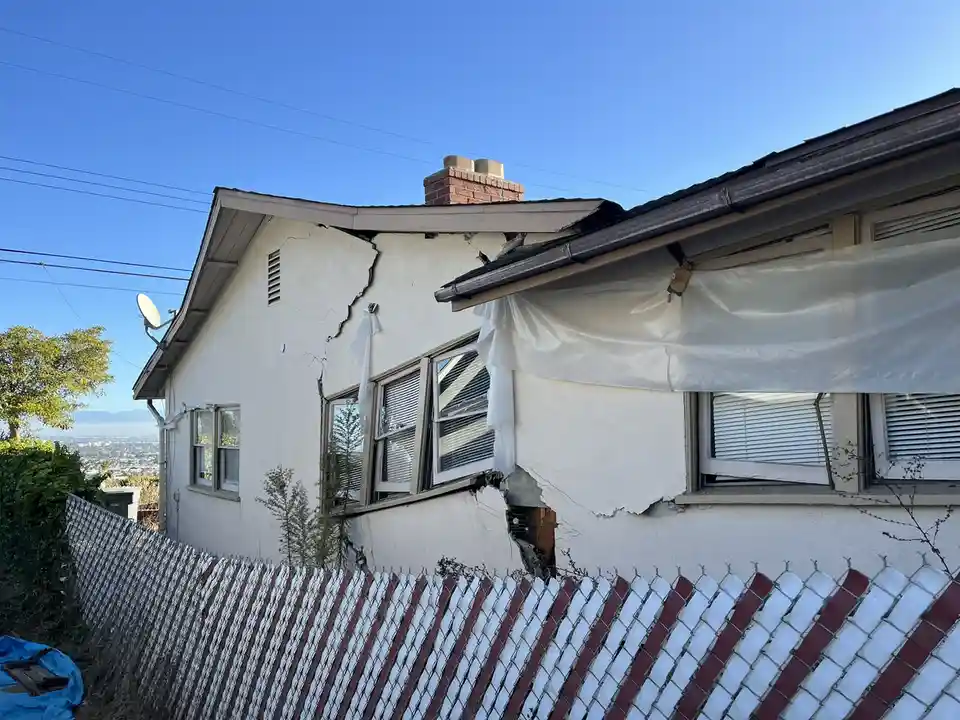 A house with severe structural damage, including a large crack and hole in the wall, a broken window, and a tarp covering…