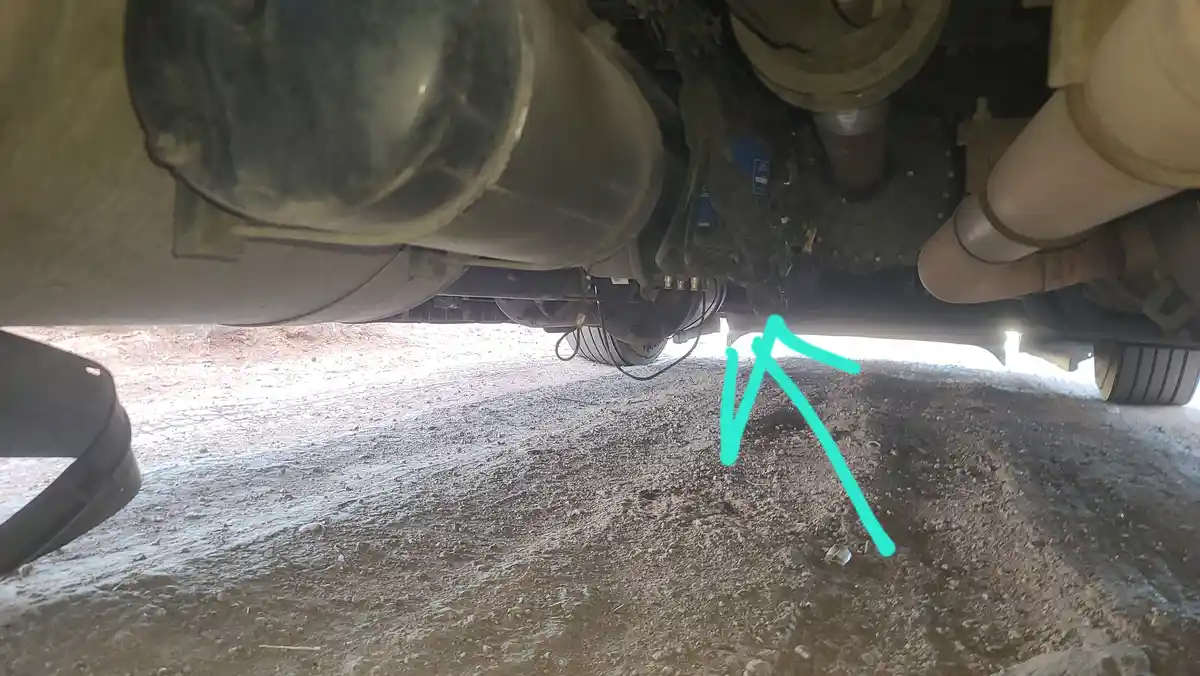 A low-angle photo looking under a vehicle at a gravelly ground surface, with a bright cyan arrow drawn on the image pointing…