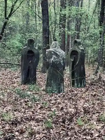 Three moss-covered, hooded stone statues stand in a forest clearing covered in fallen leaves, with trees and green foliage…