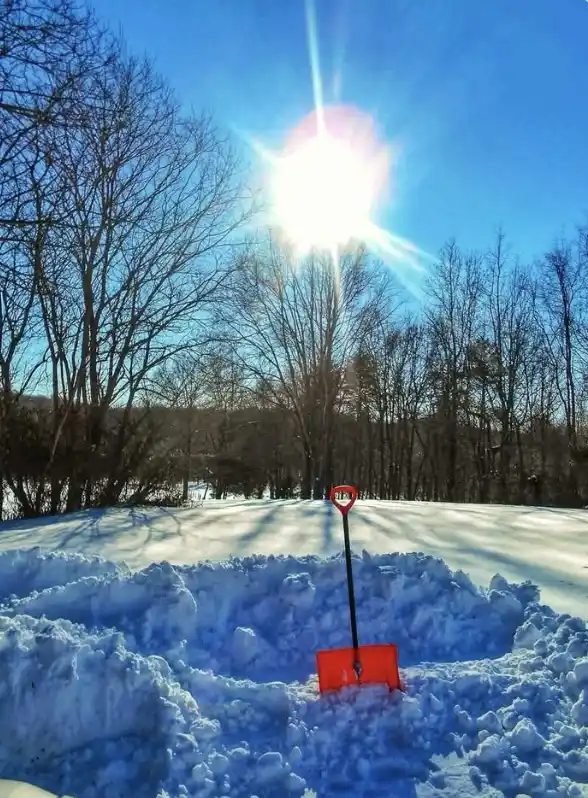 A bright orange snow shovel is stuck upright in a pile of snow, with a small red stop sign attached to its handle. The…