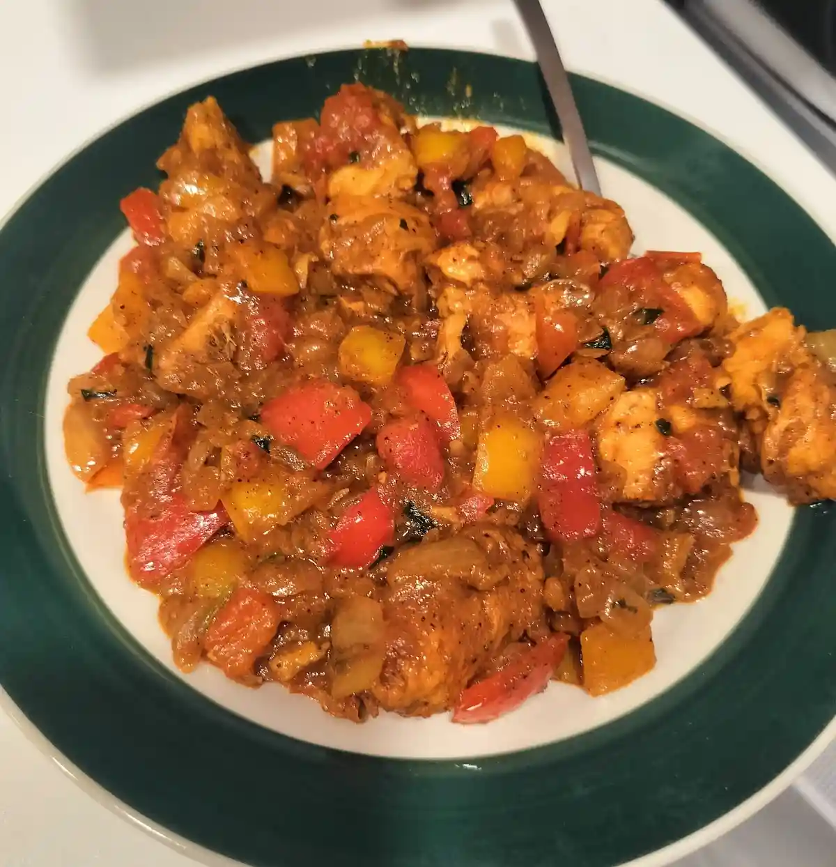 A close-up photo shows a plate of chicken and bell peppers in a thick, reddish sauce, served on a white plate with a green…