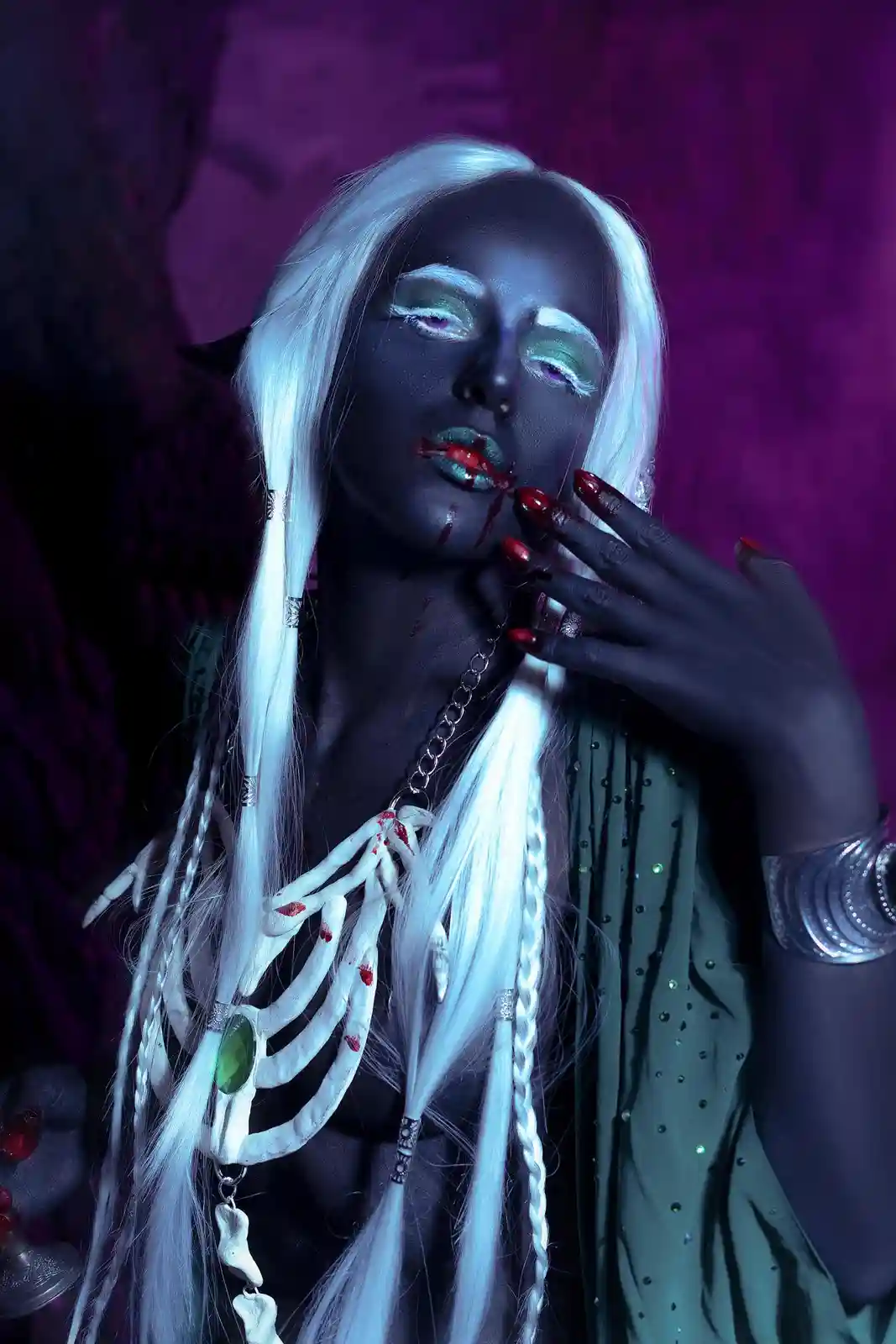 A person with dark skin, white hair, and dramatic makeup poses with blood on their lips and fingers. They wear a green…