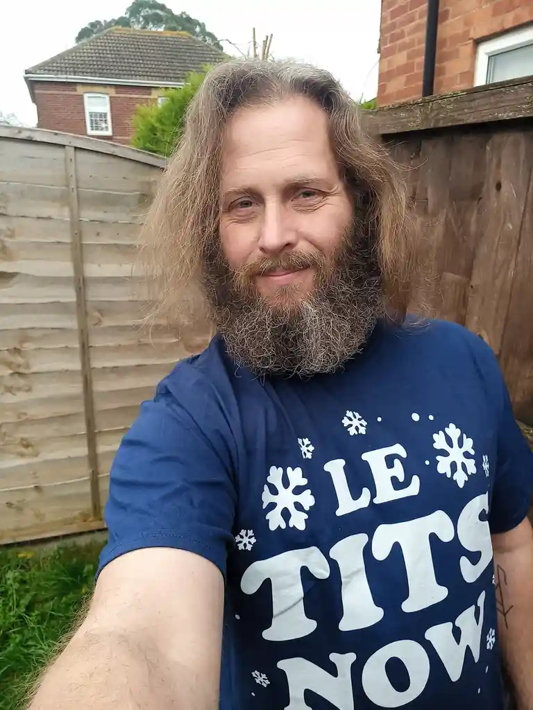 A man with long hair and a beard takes a selfie outdoors in front of a wooden fence, wearing a blue t-shirt with white…