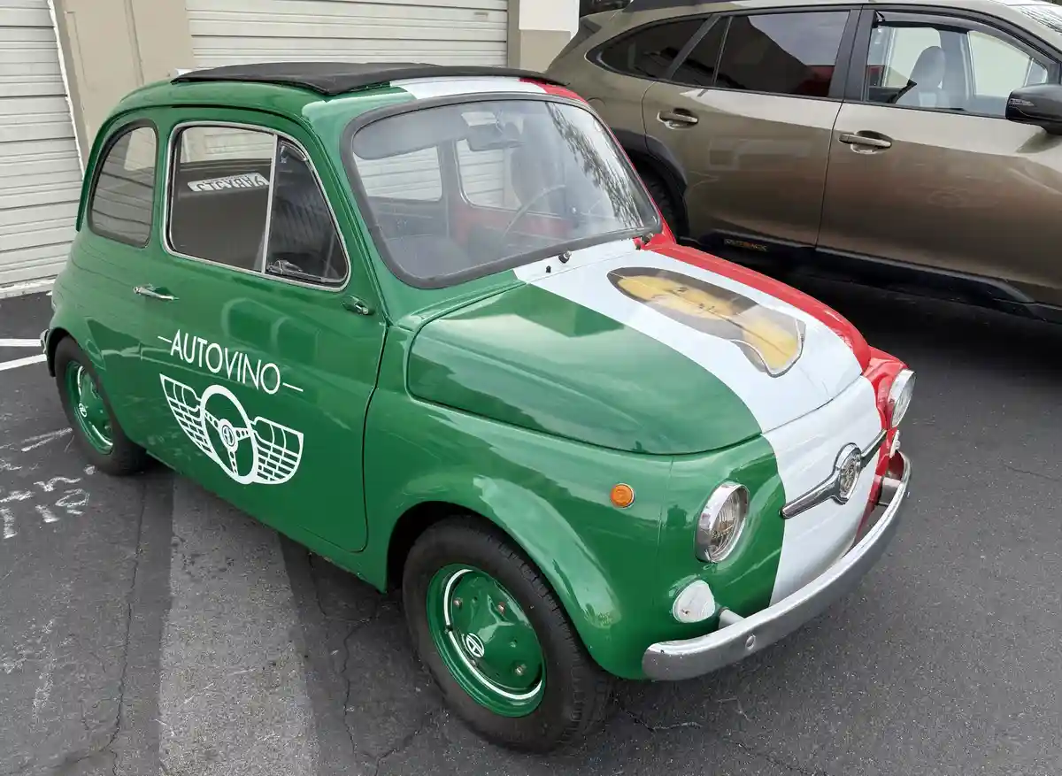 A vintage green, white, and red Fiat 500 car is parked in a lot, featuring a logo on its side that reads 'AUTOVINO' with a…