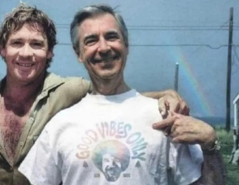 Two men are smiling and posing for a photo outdoors with a rainbow visible in the background. The man on the right, wearing…