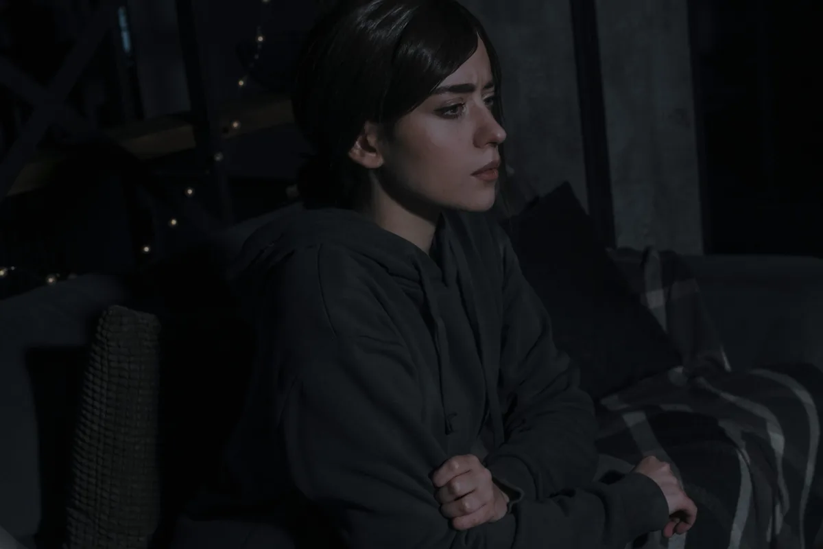 A woman with dark hair in a dark hoodie sits on a couch in a dimly lit room, looking off to the side with a pensive or sad…