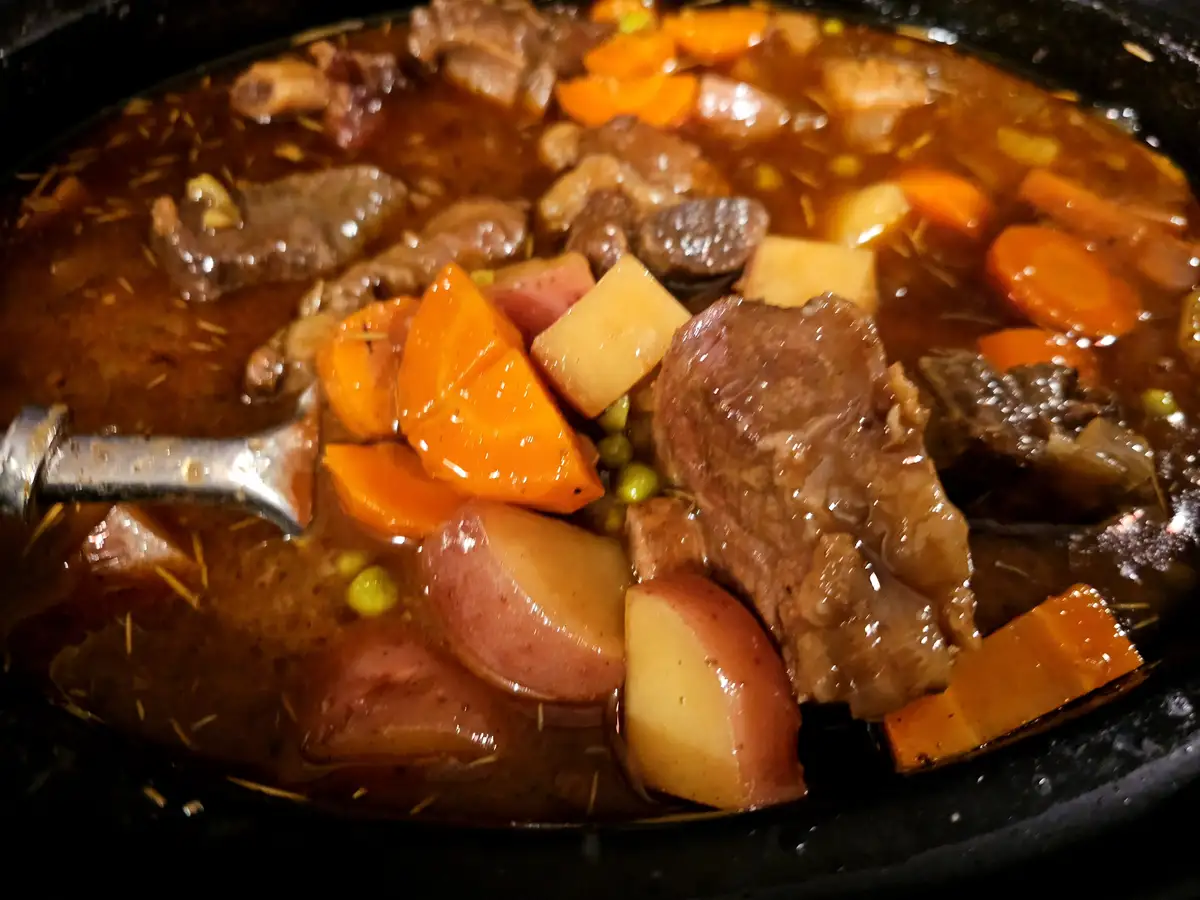 A close-up photo of a slow-cooked stew in a black pot, featuring chunks of beef, sliced carrots, diced potatoes, and green…
