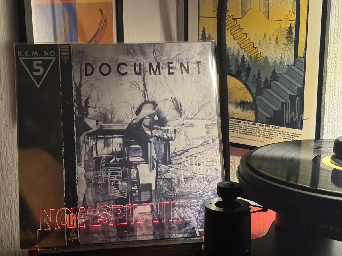 A vinyl record sleeve for R.E.M.'s album 'Document' is propped up next to a turntable with a record on it. The album cover…