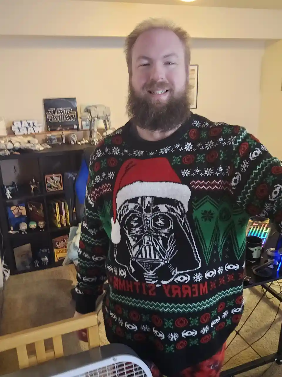 A man with a beard and blonde hair smiles while wearing a black Christmas sweater featuring Darth Vader wearing a Santa hat…