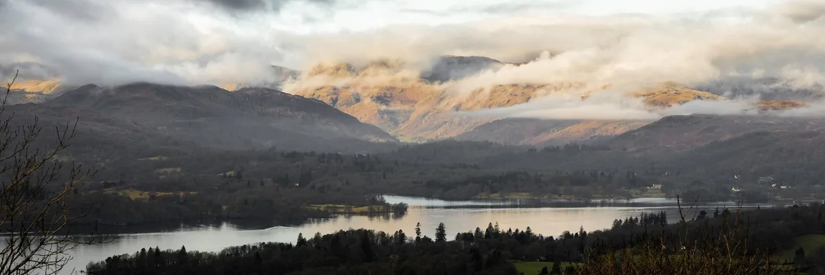 A scenic landscape photo of a lake surrounded by rolling hills and mountains partially obscured by clouds, with bare tree…